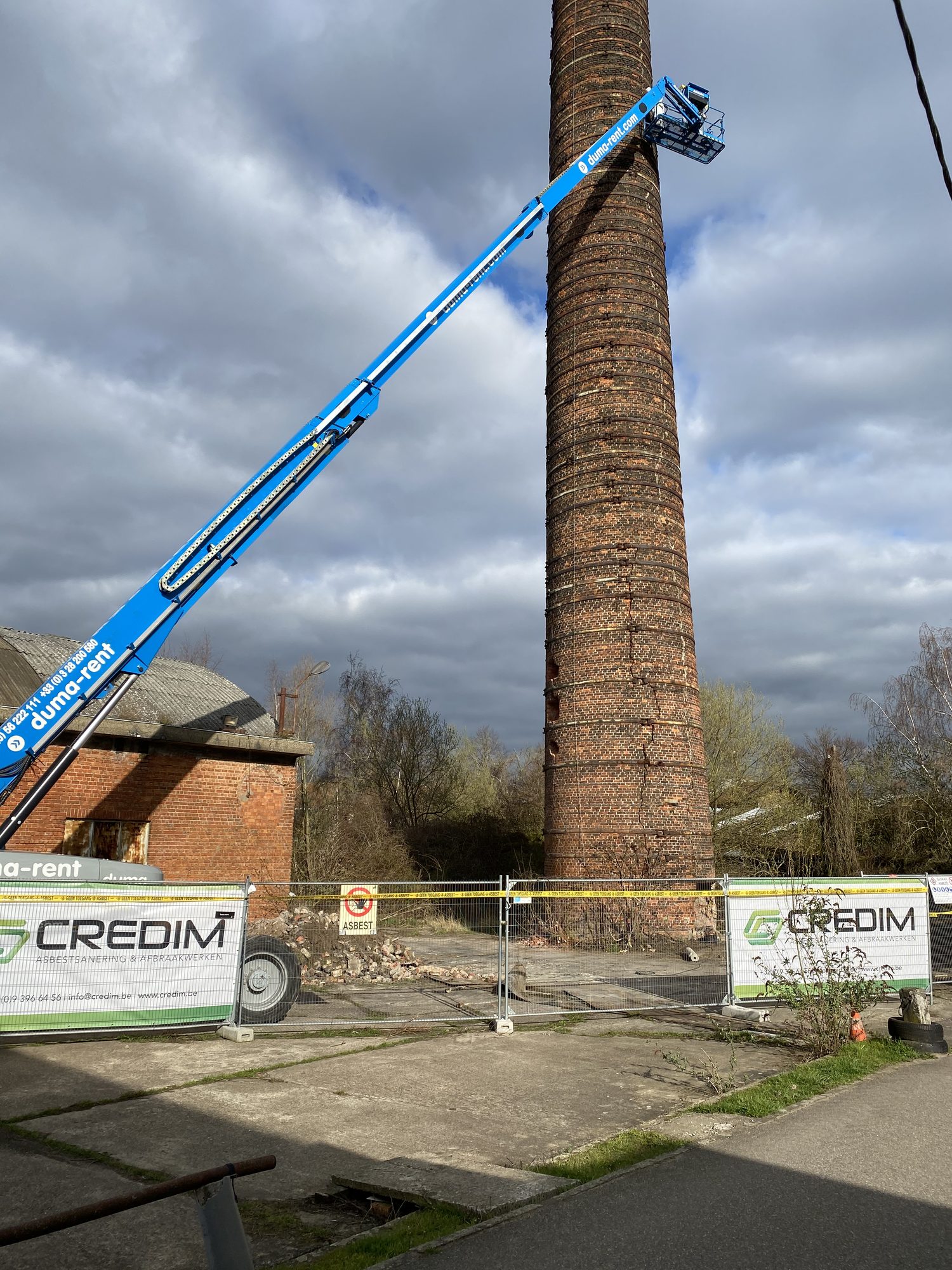 Nacelle élévatrice près d'une cheminée d'usine en briques pour le retrait de mastic, avec bâches de chantier Credim