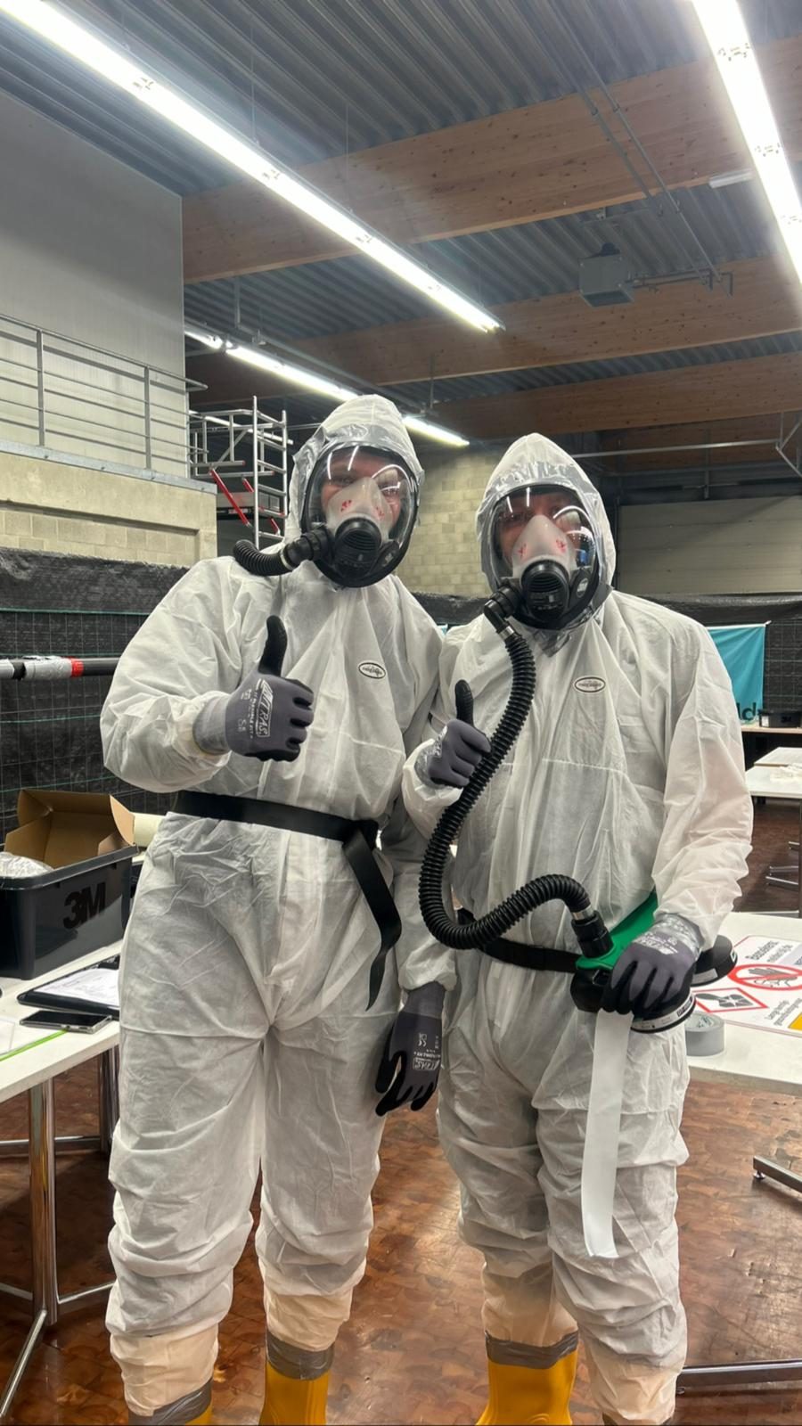 Two people in disposable coveralls with full-face masks and breathing hoses