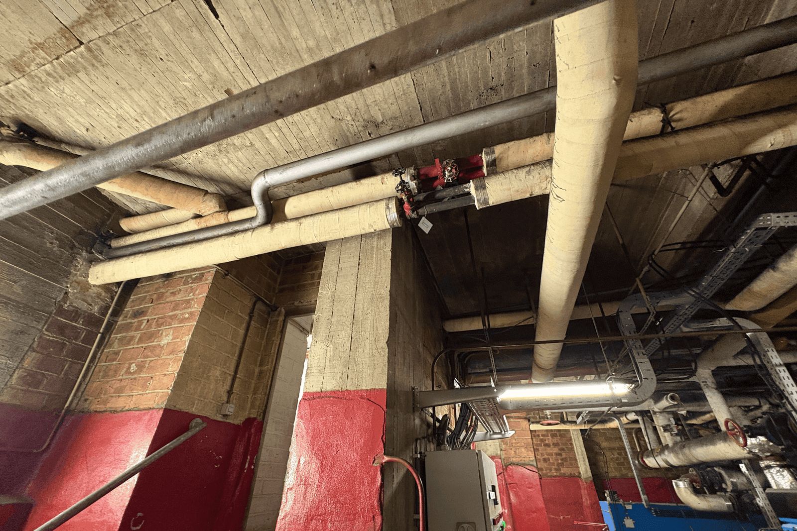 Many insulated pipes and valves along the ceiling of a room with brick walls