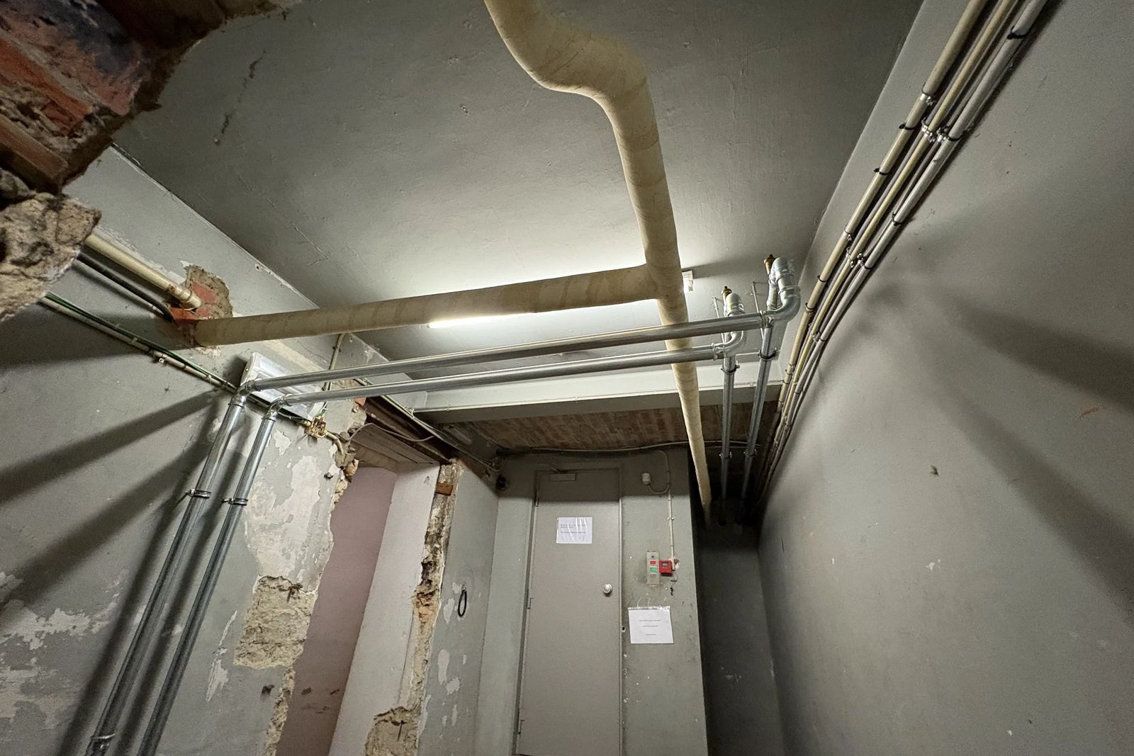 Insulated pipe along the ceiling, with metal pipes and damaged plaster on the walls