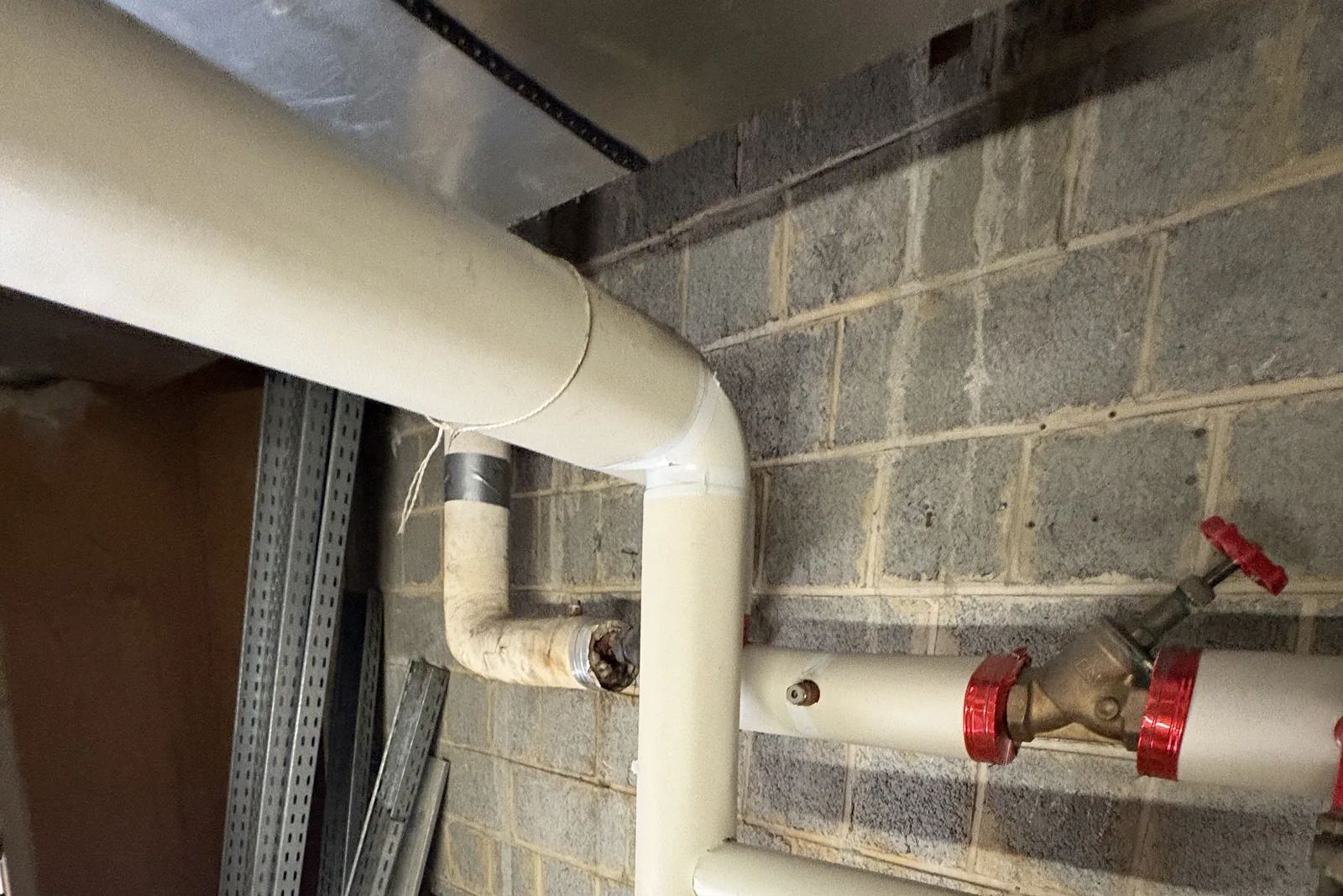 White insulated pipe with a bend along a concrete block wall, and a valve with a red handwheel