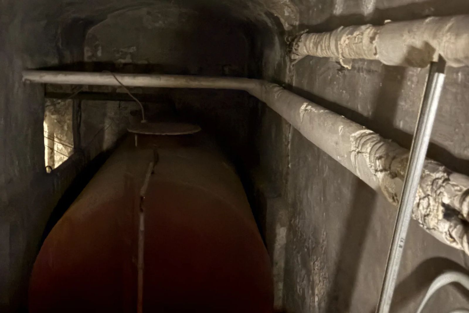 Espace étroit et sombre avec une grande cuve et des conduites isolées abîmées le long du mur