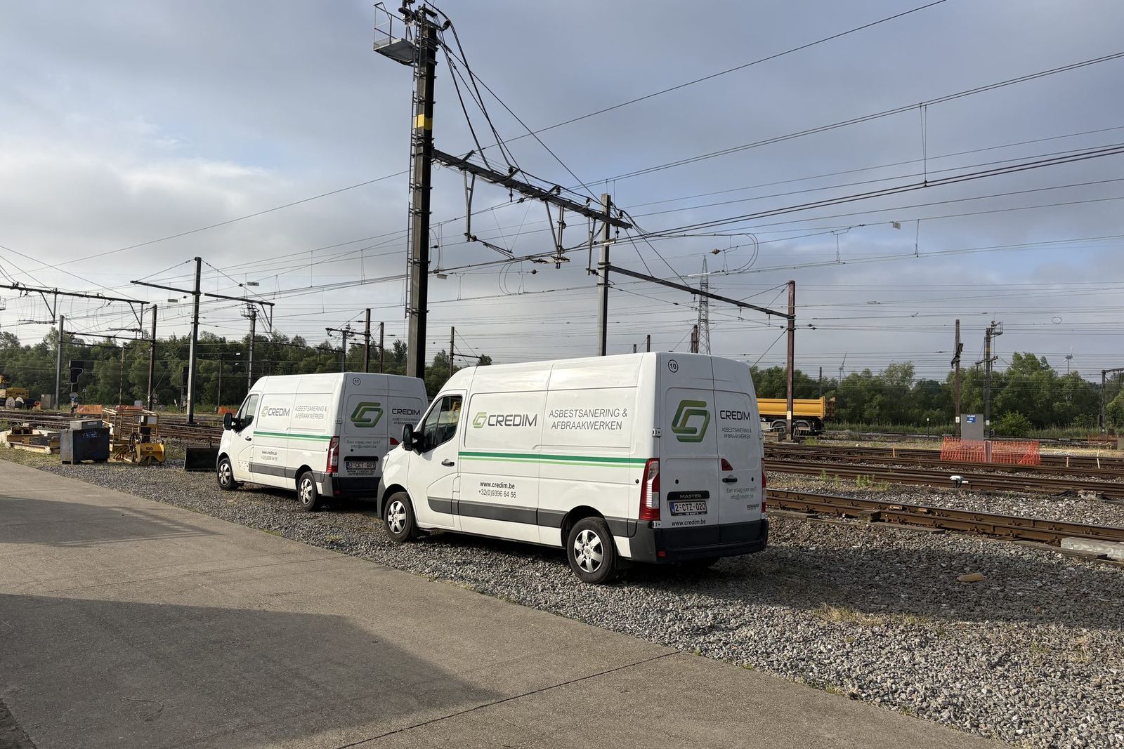 Twee bestelwagens van Credim geparkeerd op grind naast spoorlijnen met bovenleiding