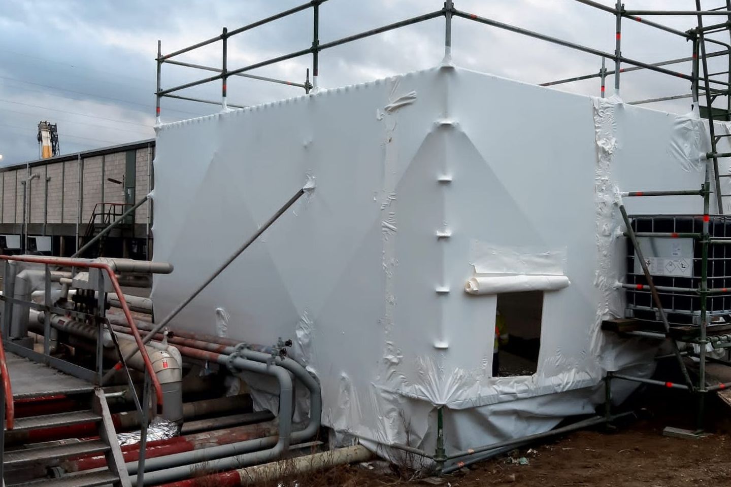 Zone hermétique en film blanc sur échafaudage, sur un site industriel, avec conduites et escalier au premier plan
