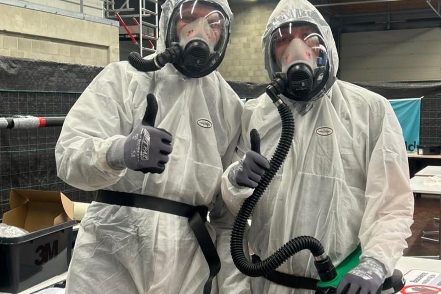 Two people in white disposable coveralls and full-face masks with hoses, giving a thumbs up
