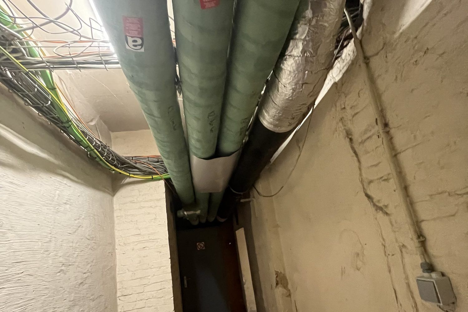 Narrow corridor with green insulated pipes bearing an asbestos label along the ceiling
