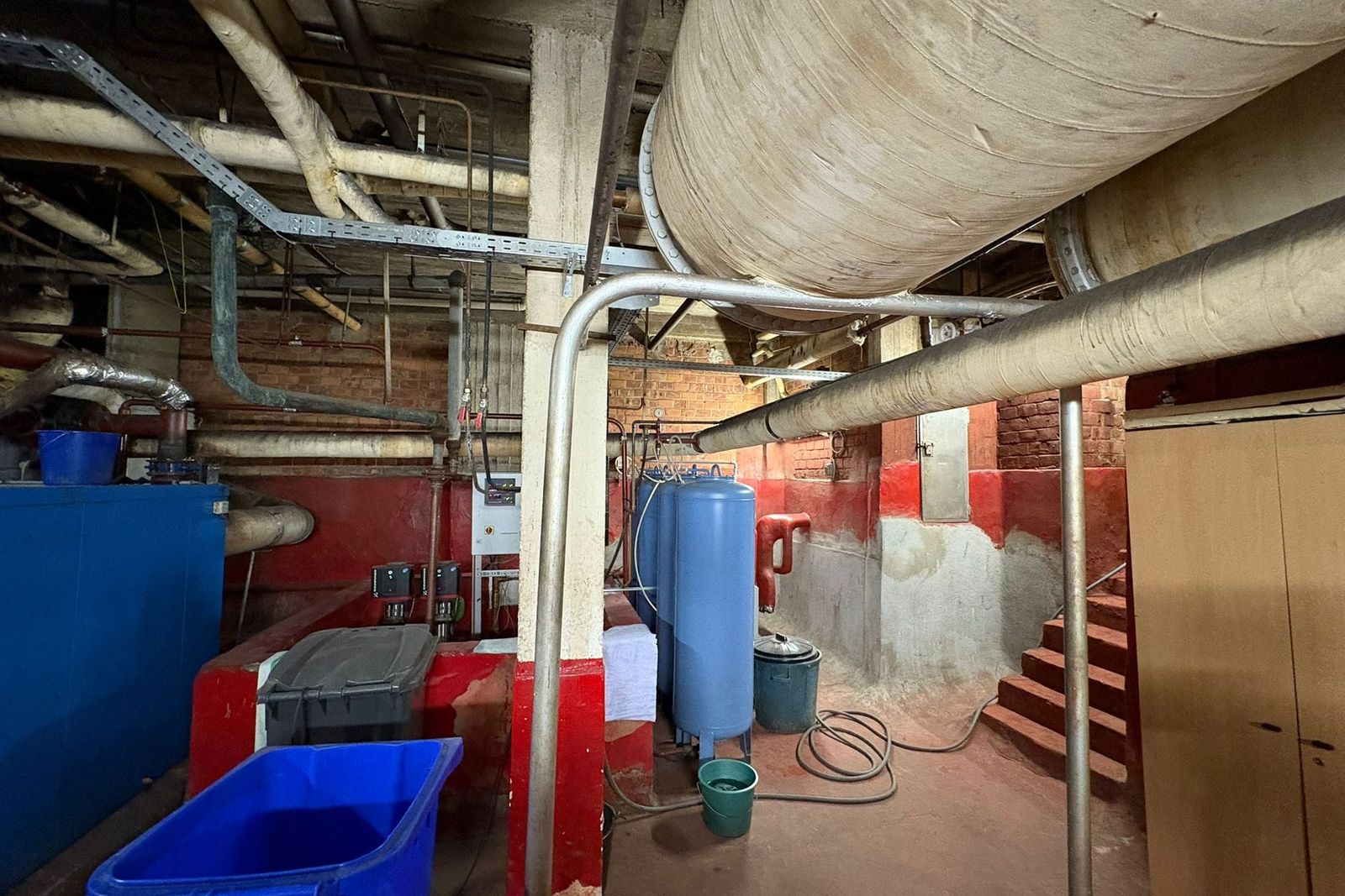 Ruimte met dikke leidingisolatie aan het plafond, blauwe vaten en deels rood geschilderde muren