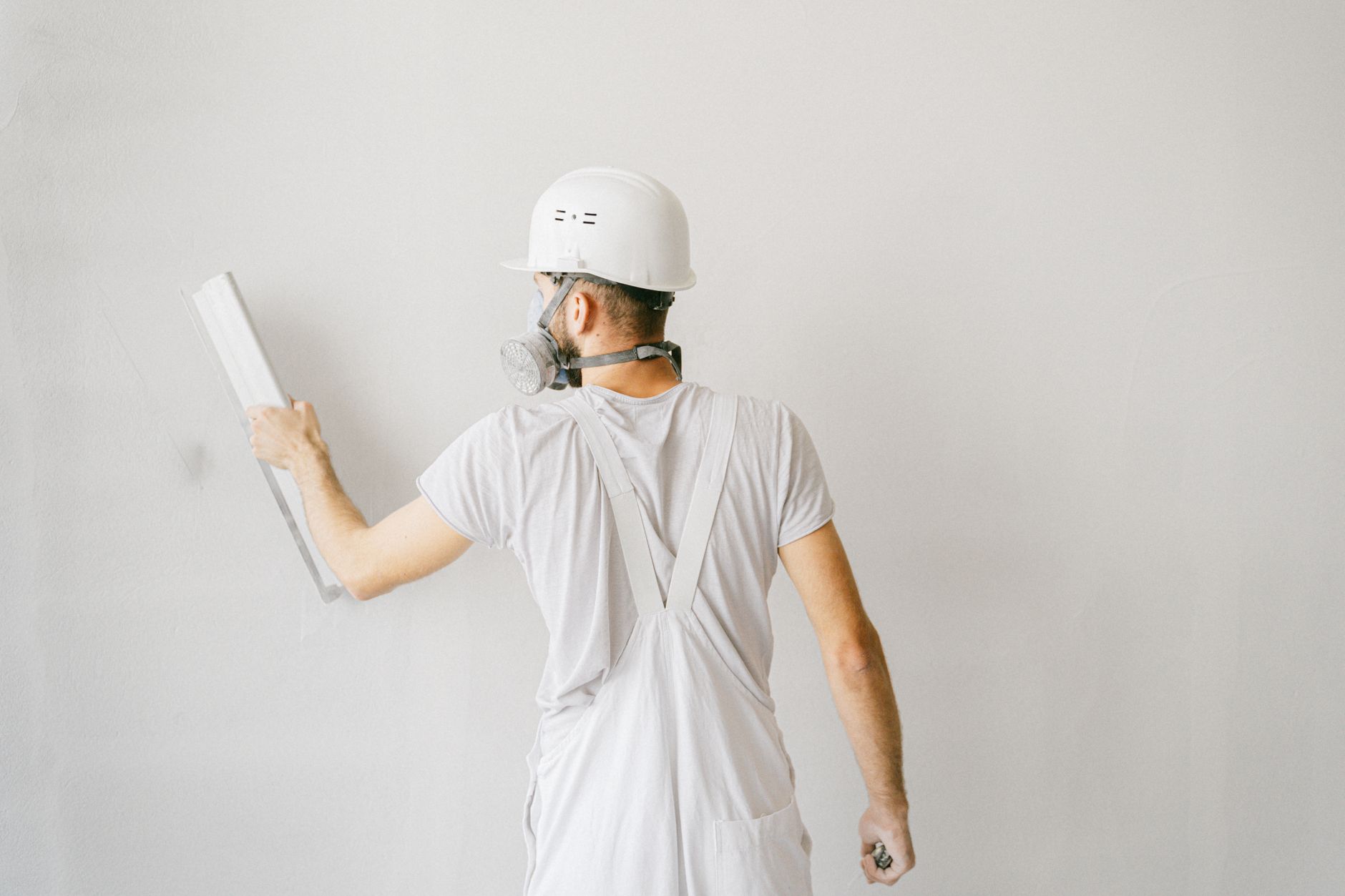Man in witte overall met helm en ademmasker houdt een lange afwerkspaan tegen een lichtgrijze muur